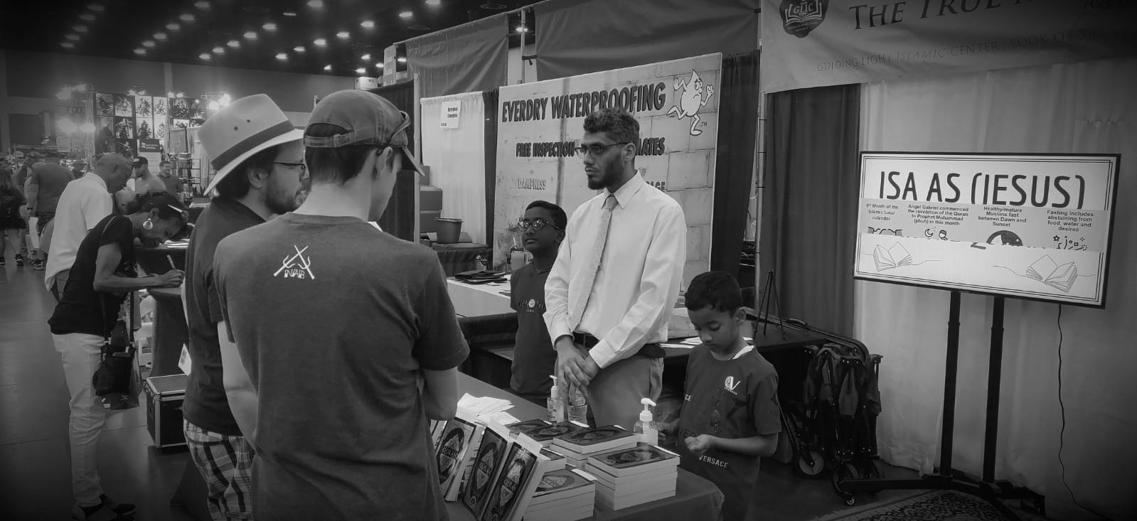 A Guiding Light volunteer and two boys talking with visitors at an outreach booth stacked with Qurans